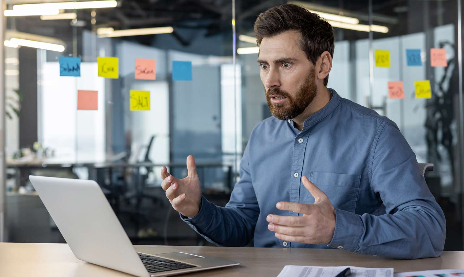 How to Avoid Digital Marketing Panic 1 A man sitting at a desk in front of a laptop gestures with his hands and looks surprised. Colorful sticky notes are visible on a glass wall behind him.